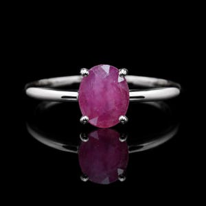 Può includere: Un anello color argento con una gemma centrale dal taglio ovale di colore rosso rosato intenso, fissata da una montatura a quattro griffe. Il gambo è liscio e lucido, riflette la luce su uno sfondo scuro, esaltando la chiarezza della pietra.
