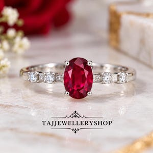 May include: A silver-toned ring featuring a large, oval-cut deep red gemstone at the centre, flanked by two small, round, clear stones on each side of the band. The text TAJJEWELLERYSHOP is printed in black below the ring on the surface.