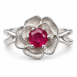 Puede incluir: Un anillo de plata con una piedra preciosa roja vibrante de corte redondo en el centro de un diseño floral texturizado y superpuesto. Los pétalos tienen un acabado mate y grabado, mientras que la banda es lisa, pulida y se divide en dos.