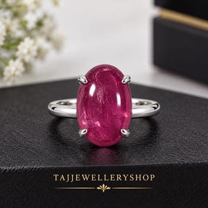 May include: A silver ring featuring a large, oval-shaped, deep pink cabochon gemstone held by four prongs. The band is smooth and polished. The text TAJJEWELLERYSHOP is printed in gold at the bottom of the image with a decorative emblem.