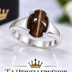 Puede incluir: Un anillo de plata con una piedra preciosa marrón ovalada con efecto ojo de gato, sujeta por cuatro garras. El aro es liso y metálico. Debajo del anillo, aparece el texto TAJJEWELLERYSHOP con un icono de corona dorada sobre la palabra TAJ.