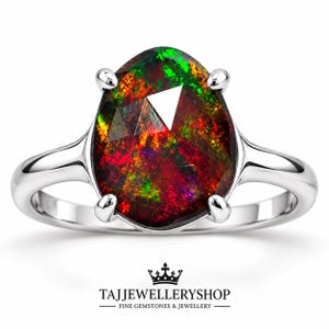 May include: A silver ring featuring a large, pear-shaped black opal with vibrant red and green flashes of color. The gemstone is held by four prongs on a smooth, polished silver band. Below the ring is the text TAJJEWELLERYSHOP FINE GEMSTONES & JEWELLERY.