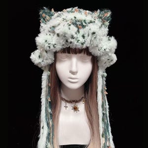 Peut inclure: Un chapeau en fausse fourrure blanche avec des protubérances en forme d'oreilles, orné de détails floraux verts et pêche. De longues mèches tressées avec des ornements similaires pendent. Le chapeau est présenté sur une tête de mannequin.