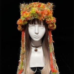 May include: A colorful, furry hat with ear-like protrusions, featuring shades of orange, yellow, and green. Long, multi-colored strands hang down, complementing the hat's vibrant design. The hat is displayed on a mannequin head.