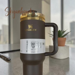 Puede incluir: Un vaso Stanley Quencher H2.0 de color marr&oacute;n oscuro con una capacidad de 1,18 L. El vaso cuenta con un asa a juego, una tapa de tono dorado y una etiqueta blanca con el texto The Flowstate Quencher H2.0 Tumbler 40 oz 1.18 L All Day Hydration.