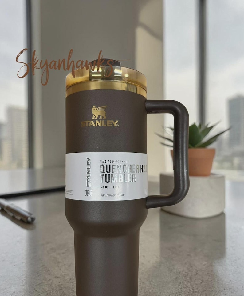 Puede incluir: Un vaso Stanley Quencher H2.0 de color marr&oacute;n oscuro con una capacidad de 1,18 L. El vaso cuenta con un asa a juego, una tapa de tono dorado y una etiqueta blanca con el texto The Flowstate Quencher H2.0 Tumbler 40 oz 1.18 L All Day Hydration.