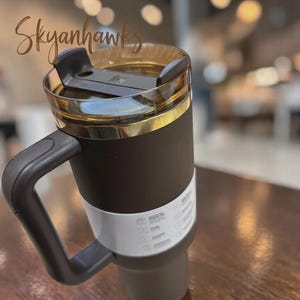 Puede incluir: Un vaso t&eacute;rmico de color marr&oacute;n oscuro con un asa resistente y una tapa transparente de tono &aacute;mbar con cierre deslizante negro. Una banda de papel blanca con texto e iconos impresos envuelve la secci&oacute;n inferior del recipiente mate.