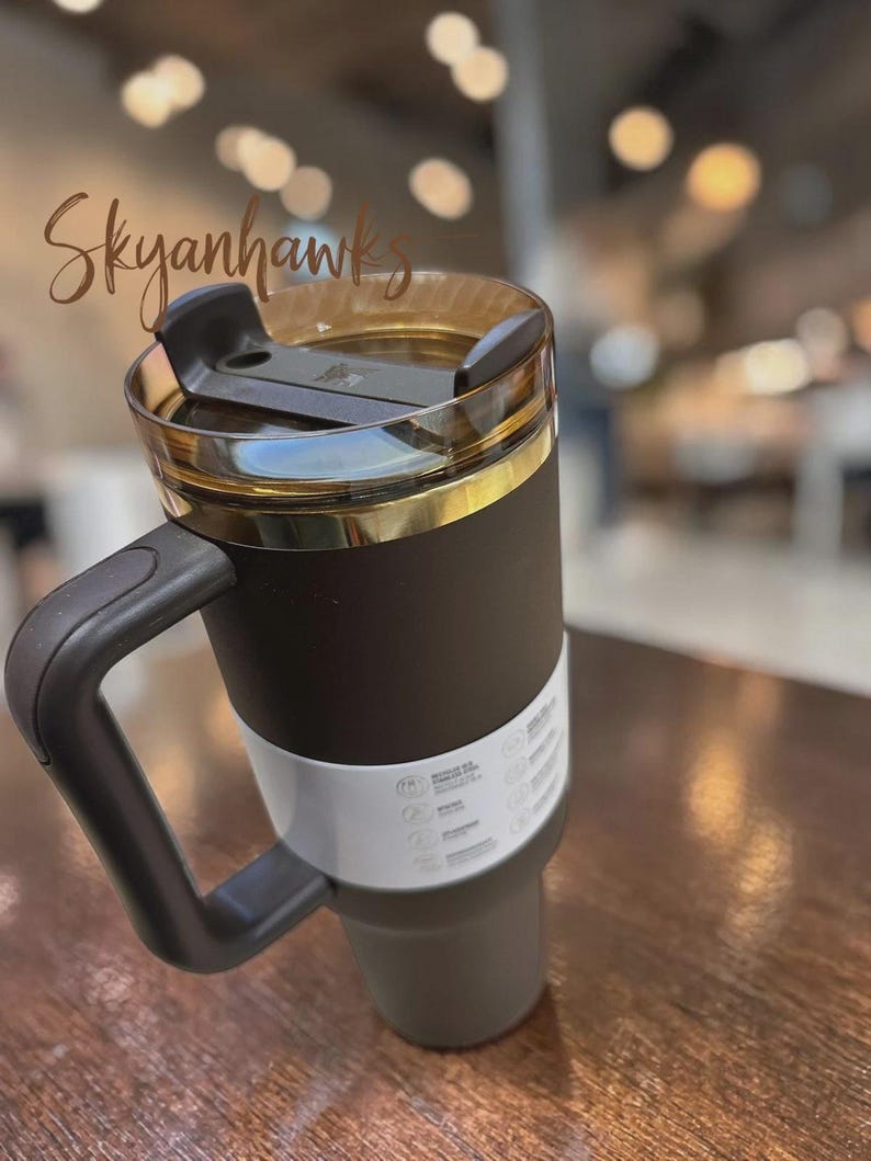 Puede incluir: Un vaso t&eacute;rmico de color marr&oacute;n oscuro con un asa resistente y una tapa transparente de tono &aacute;mbar con cierre deslizante negro. Una banda de papel blanca con texto e iconos impresos envuelve la secci&oacute;n inferior del recipiente mate.