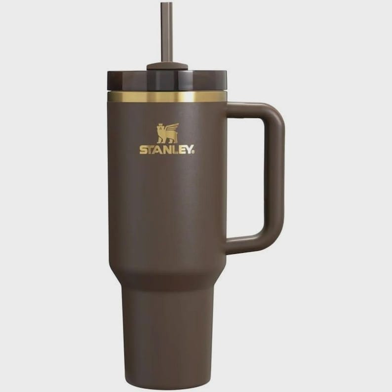 Puede incluir: Un vaso t&eacute;rmico de color marr&oacute;n mate con asa a juego y tapa marr&oacute;n oscuro que incluye una pajita central. El cuerpo muestra el logotipo de Stanley en dorado con un emblema de oso alado. El dise&ntilde;o incluye una banda dorada bajo la tapa.