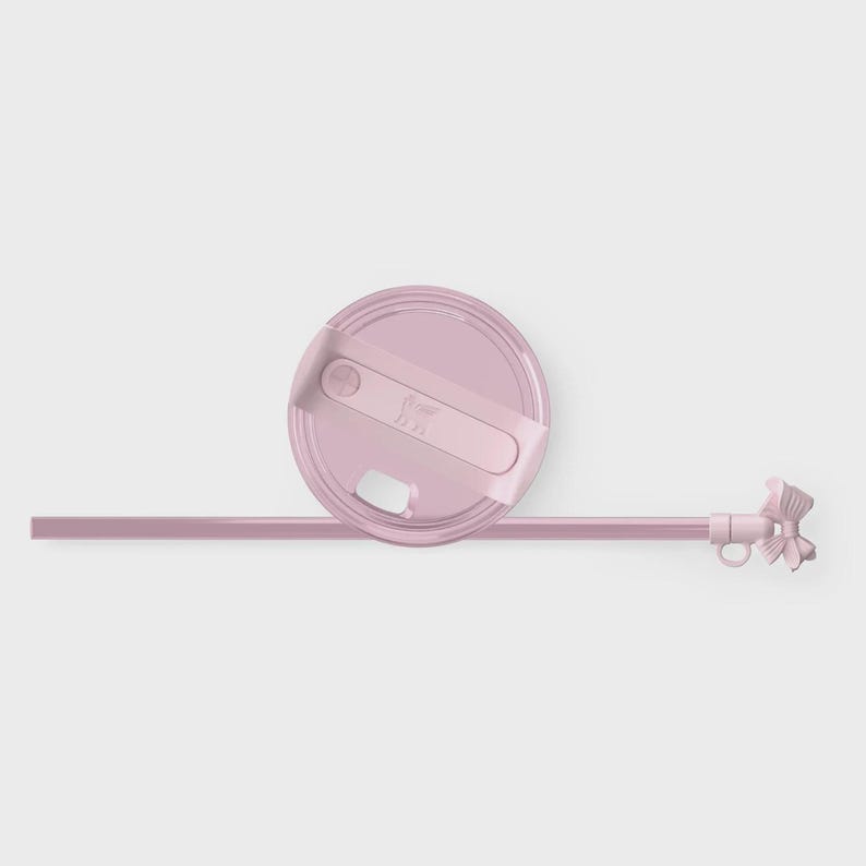 May include: A light pink circular tumbler lid with a rotating closure mechanism and a matching light pink straw. The straw features a small, decorative bow-shaped attachment at the top end, providing a distinct accent to the drinkware accessory.
