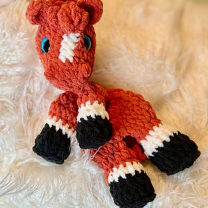 Peut inclure: Un jouet de cheval au crochet fait à la main, aux tons orange, blanc et noir. Le cheval a des yeux bleus et une marque blanche sur le front. Le jouet est posé sur une surface blanche et duveteuse.
