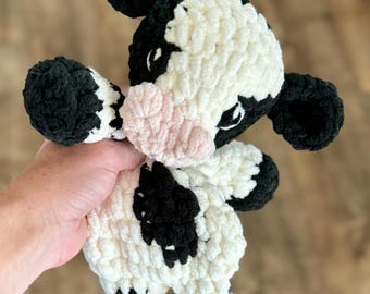 May include: A handmade plush cow toy with a black and white pattern. The cow has black ears, hooves, and patches, with a pink nose and a fluffy, textured appearance. The toy is held in a person's hand.