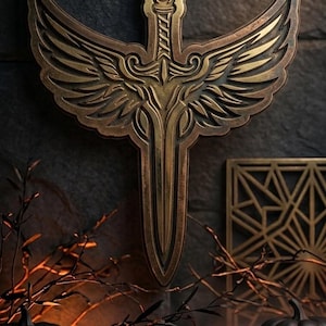 Könnte beinhalten: Eine bronzefarbene Wandplakette mit einem zentralen Schwertdesign und symmetrischen, detaillierten Flügeln, die vom Griff ausgehen. Das Stück hat ein metallisches Finish mit dunklen Vertiefungen und zeigt filigrane Gravuren.