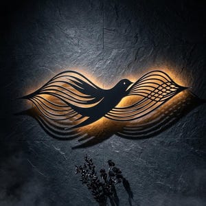 May include: A black metal wall art piece shaped like a bird in flight with elongated, flowing wing patterns. The design features a warm, golden backlighting that creates a glowing silhouette against the dark, textured wall surface behind it.
