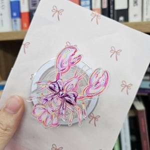 May include: A clear acrylic lobster-shaped pin with pink and purple accents, a purple bow, and glitter details. The pin is on a pink card with small bow patterns.