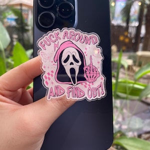 Puede incluir: Un smartphone negro con una pegatina rosa y blanca con una máscara de Scream y el texto "FUCK AROUND AND FIND OUT!". La pegatina también incluye una mano haciendo la peineta y gráficos de mariposas.