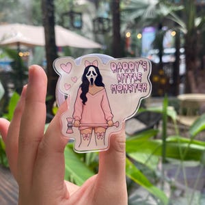 May include: A clear acrylic charm featuring a cartoon illustration of a character in a Scream mask, wearing a pink outfit and holding a double-sided axe. The text "Daddy's Little Monster" is included in the design, along with hearts and bows.