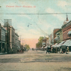 May include: A vintage postcard depicting a street scene in Kewanee, Illinois. The postcard shows a street lined with brick buildings, some with awnings. The street is empty except for a few people and horse-drawn carriages. The text "Tremont Street - Kewanee, Ill" is printed at the top of the postcard.