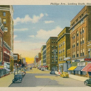 May include: Vintage postcard depicting Phillips Avenue in Sioux Falls, South Dakota, looking south. The image shows a street lined with buildings, shops, and vintage cars. The sky is a mix of blue and yellow.