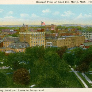 May include: Vintage postcard depicting a general view of Sault Ste. Marie, Michigan, from the Soo Locks. The Ojibway Hotel and Annex are in the foreground. The image shows a cityscape with buildings, trees, and a blue sky with clouds.