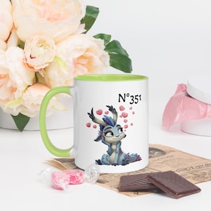 Pode incluir: Caneca de cerâmica branca com alça e borda verdes. A caneca apresenta uma ilustração de desenho animado de uma criatura azul com detalhes de coração e o número "N°351". A caneca é cercada por flores cor-de-rosa e chocolates.
