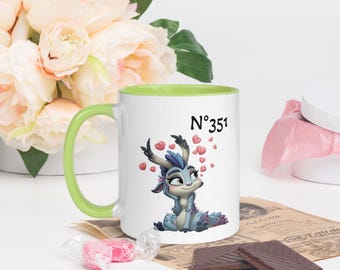 May include: White ceramic mug with a green handle and rim. The mug features a cartoon illustration of a blue creature with heart accents and the number "N°351". The mug is surrounded by pink flowers and chocolates.