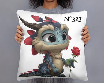 May include: White square pillow featuring a cartoon dragon holding a red rose. The dragon has blue, red, and tan coloring with large eyes. The pillow has the number "N°323" printed in black.