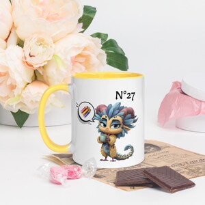 May include: White ceramic mug with a yellow handle and interior. The mug features a cartoon illustration of a mythical creature holding a cup, with the number "27" and a speech bubble with pencils. The mug is decorated with a floral arrangement.