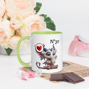 Puede incluir: Taza de cerámica blanca con asa y borde verdes, con un dragón de dibujos animados con una burbuja de pensamiento de corazón y el número 30. El dragón sostiene una taza pequeña. La taza está rodeada de flores y chocolate.