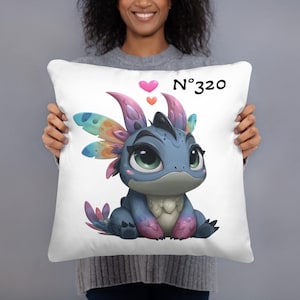 Può includere: Cuscino bianco con un'illustrazione di un drago dei cartoni animati. Il drago è blu con ali e accenti colorati e grandi occhi verdi. Il cuscino ha il testo "N°320" e due cuori.