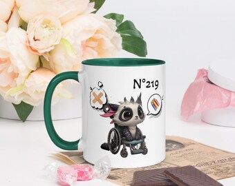 May include: White ceramic mug with a green interior and handle. The mug features a cartoon illustration of a creature in a wheelchair, with the number "219" printed on the side. The creature has large eyes and is surrounded by thought bubbles.