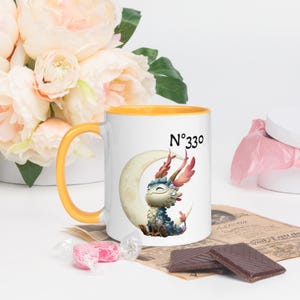 Puede incluir: Taza de cerámica blanca con asa y borde amarillos, con un personaje de dragón de dibujos animados sentado en una luna creciente. La taza tiene el número "N°330" impreso. La taza está decorada con un diseño de dragón colorido.