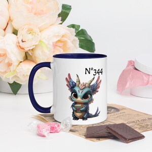 Könnte beinhalten: Weißer Keramikbecher mit blauem Innenraum und Griff. Der Becher zeigt eine Cartoon-Drachen-Illustration mit blauen, roten und braunen Farben. Der Text "N°344" ist über dem Drachen aufgedruckt. Der Becher ist mit einem Blumenarrangement und Pralinen dekoriert.