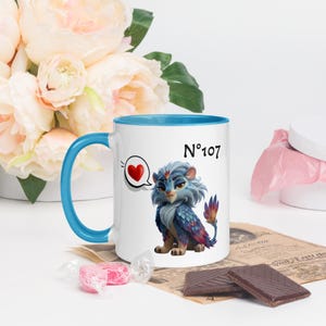 Puede incluir: Taza de cerámica blanca con interior y asa azules. La taza presenta una ilustración de dibujos animados de una criatura azul y morada con un bocadillo de corazón y el número "107".
