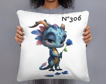 May include: White throw pillow featuring a cartoon dragon holding a blue rose. The dragon is blue with pink and blue accents, and has curved horns. The pillow has the text "N°306" in black.