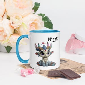 Puede incluir: Taza de cerámica blanca con interior y asa azules, con una ilustración de un dragón de dibujos animados. La taza tiene el número "338" y pequeños corazones. La taza está rodeada de flores y chocolate.