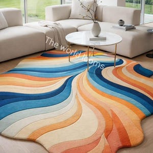 May include: A colorful, abstract area rug with a wave-like design. The rug features a blend of blue, orange, and cream hues. It is placed on a hardwood floor in a modern living room setting, with a white sofa and coffee table.