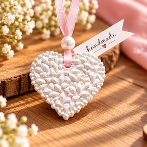May include: A white heart-shaped ornament with a textured, faceted surface. It hangs from a light pink ribbon accented by a single white pearl bead. A small white paper tag attached to the ribbon features the text handmade with a small pink heart.