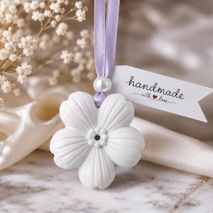 May include: A white ceramic flower ornament suspended by a light purple ribbon with a single white pearl bead. A white paper tag attached to the ribbon features the black printed text "handmade with love" with a small red heart icon included.