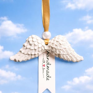 May include: White angel wings ornament with a gold ribbon and a pearl bead. A tag with the words "handmade with love" and a red heart hangs below the wings. The background is a blue sky with white clouds.