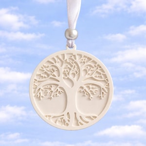 Puede incluir: Adorno circular color crema con un diseño detallado de árbol de la vida. Cuelga de una cinta blanca y una pequeña perla. El fondo es un cielo azul brillante con nubes blancas esponjosas.