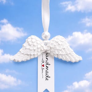 Könnte beinhalten: Weißer Engelsflügel-Anhänger mit Perle und weißem Band zum Aufhängen. Der Anhänger trägt den Schriftzug "handmade with love" auf einem weißen Anhänger mit rotem Herz. Der Hintergrund ist ein blauer Himmel mit Wolken.