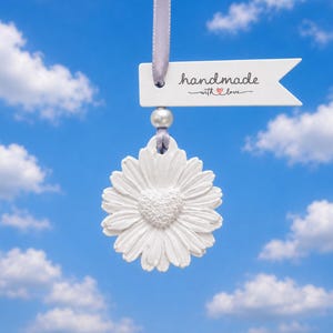 Op de afbeelding: Een wit madeliefvormig ornament met een hartvormig centrum, opgehangen aan een lichtpaars lint. Boven de bloem zit een kleine witte kraal. Een wit rechthoekig label met een puntig uiteinde bevat de tekst "handmade with love" in het zwart.