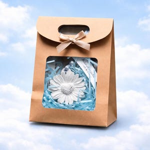 May include: A brown paper gift bag with a window, tied with a beige ribbon. Inside, a white daisy-shaped ornament rests on blue paper shreds. A small tag is visible.