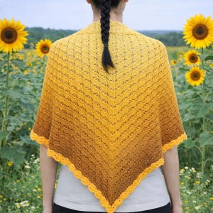 May include: A yellow and brown crocheted shawl with a scalloped edge. The shawl is worn over a white top and black trousers, with sunflowers in the background. The shawl's colours transition from yellow to brown.
