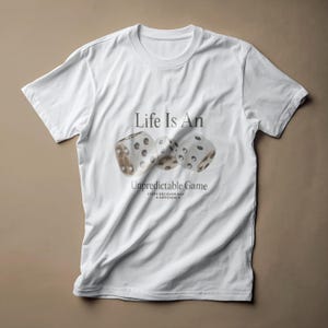 以下が含まれることがあります： 「Life Is An Unpredictable Game」の文字と3つのサイコロのイラストが描かれた白いTシャツ。サイコロは白地に茶色の影と黒い点があります。Tシャツはクルーネックです。