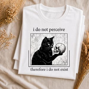 May include: White t-shirt featuring a black cat holding a skull, with the text "i do not perceive therefore i do not exist." The cat illustration is within a black rectangular frame. The shirt is folded and resting on a neutral-colored surface.