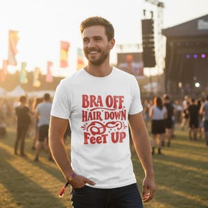 May include: White t-shirt with the red text "BRA OFF, HAIR DOWN, FEET UP." The shirt is being worn by a person at an outdoor event. The background is blurred, with people and a stage visible.