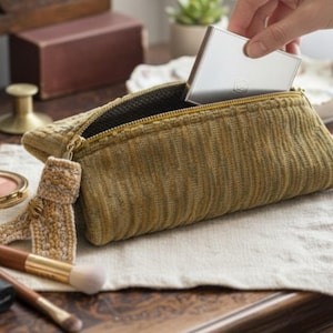 Può includere: Un beauty case in velluto a coste verde oliva e beige, con cerniera e nappa dorate. Un compatto argento viene inserito all'interno. Pennelli per il trucco e un compatto sono visibili in primo piano.