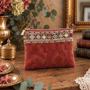 Può includere: Una pochette rettangolare in velluto color ruggine con un motivo floreale testurizzato. La parte superiore è decorata con una fascia di paillettes dorate, ricami rosa e verdi e piccoli specchietti incastonati, rifinita con una zip dorata.